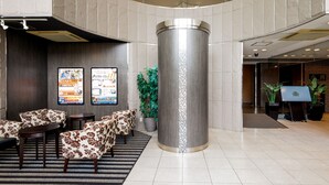 Un hall d’entrée doté d’une colonne cylindrique centrale, d’un espace salon avec des canapés à motifs et de deux écrans numériques affichant des informations.