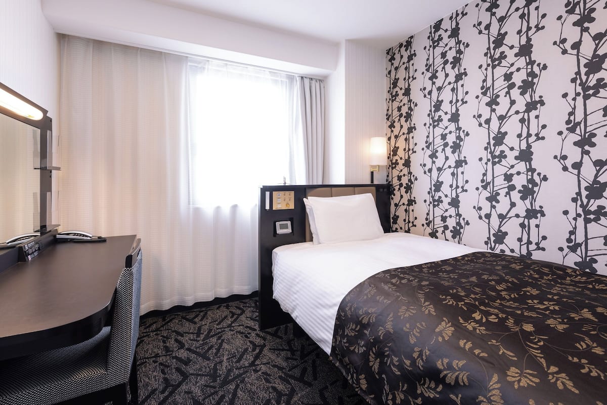 Ein Hotelzimmer mit Bett, Schreibtisch, Sessel und einem Fenster mit Vorhängen. Eine Wand zeigt ein Blumenmuster.