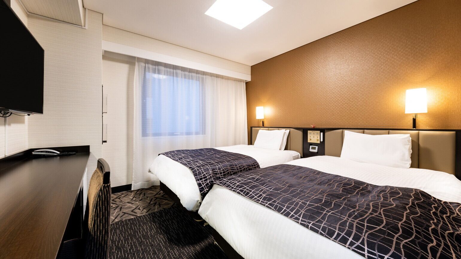 A hotel room with two beds, a desk, a TV, and a window with curtains.