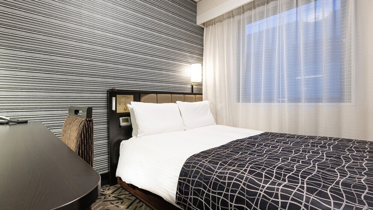 A hotel room with a bed, a desk, a wall with a striped pattern, and a window with sheer curtains.
