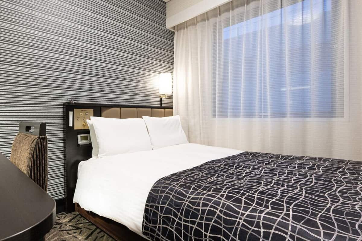 Une chambre d’hôtel avec un lit, un bureau, un mur à motifs rayés et une fenêtre avec des rideaux légers.