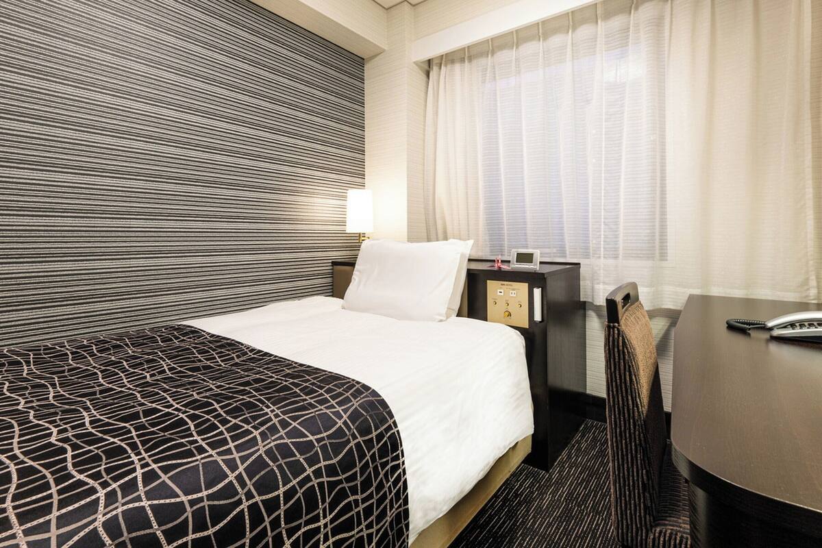 A hotel room with a bed, a desk, a chair, a nightstand, and a window with curtains.