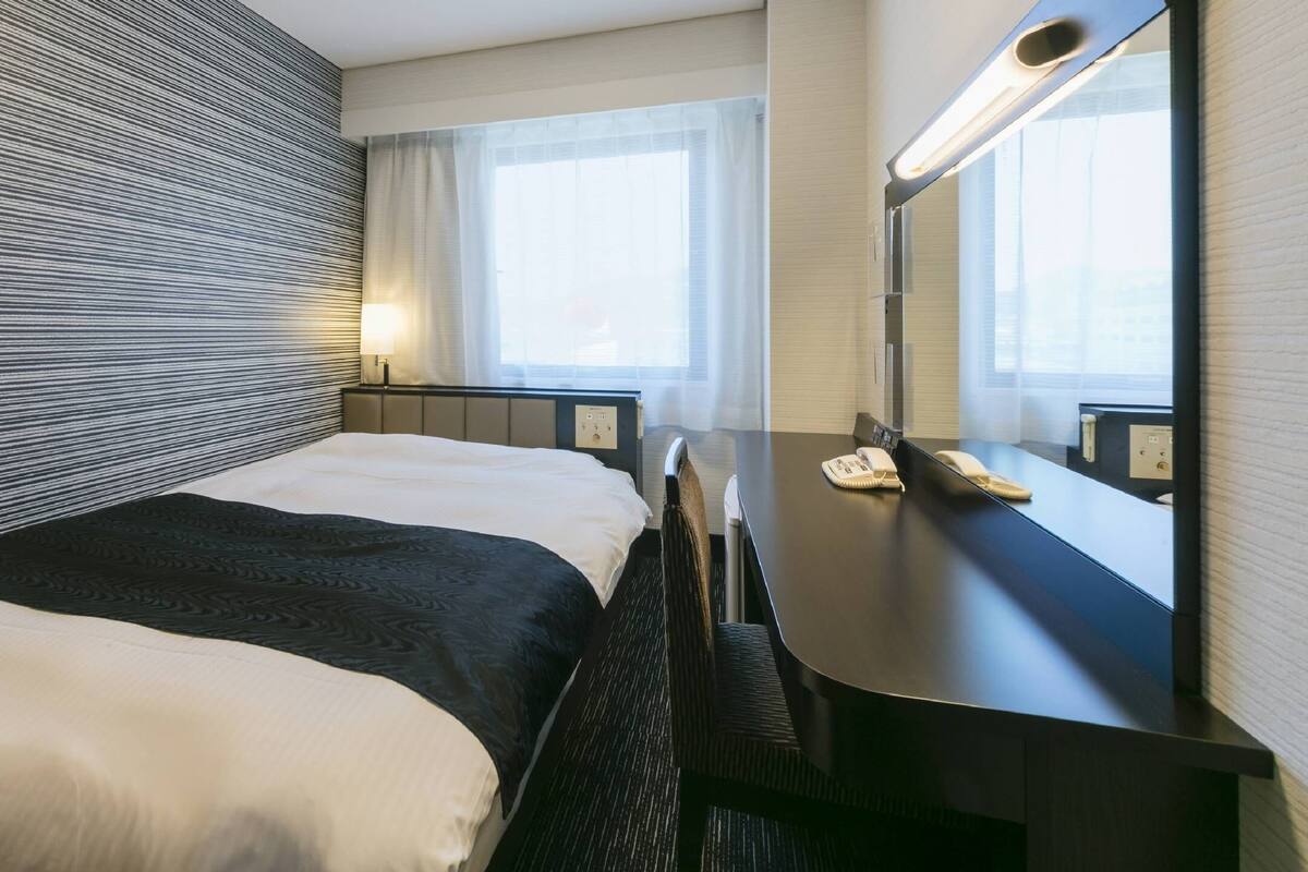 A hotel room with a bed, a desk, a mirror, and a window.
