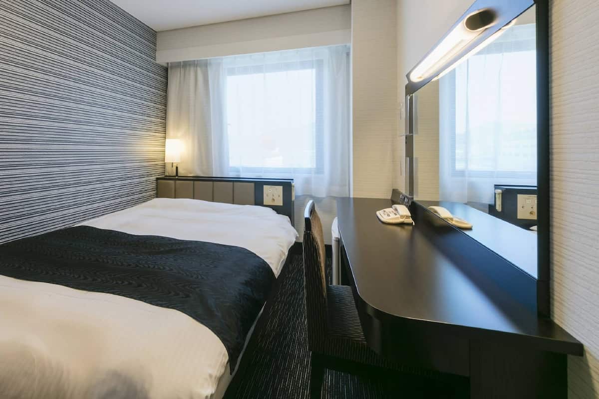 A hotel room with a bed, a desk, a mirror, and a window.