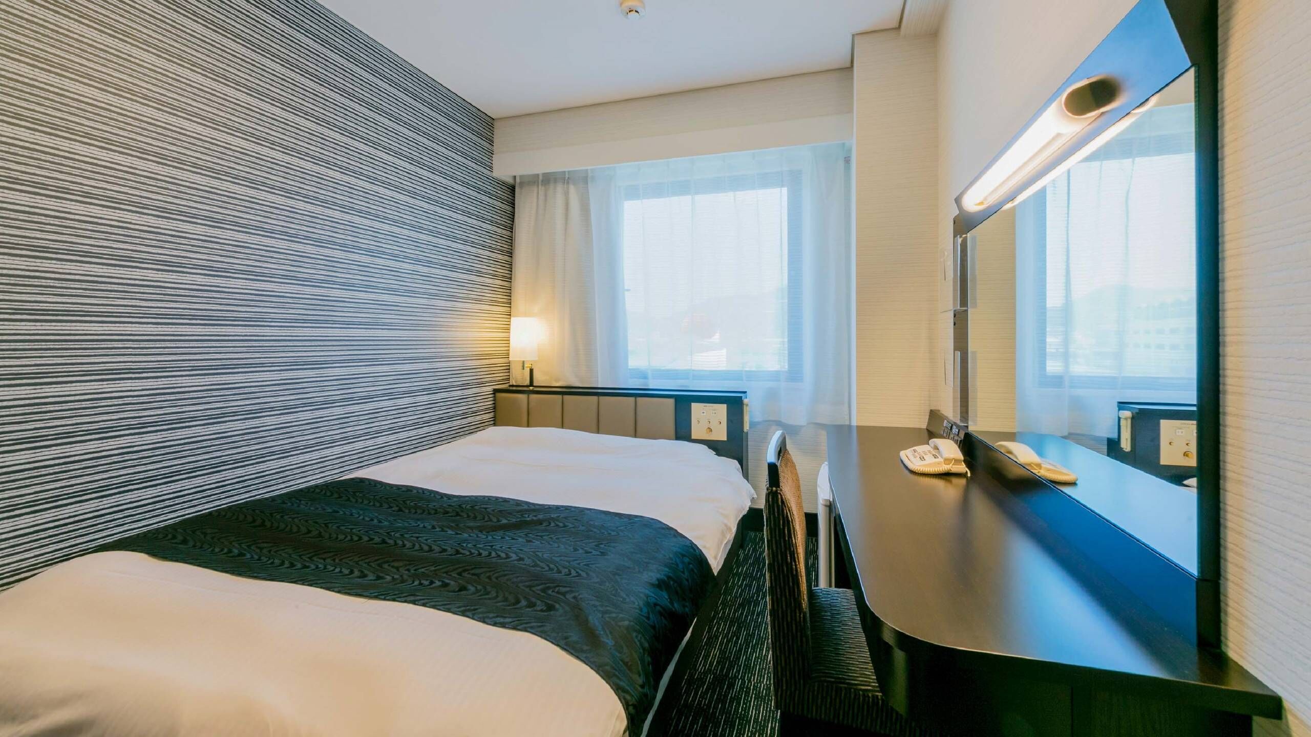 A hotel room with a bed, bedside table, lamp, and a large mirror.
