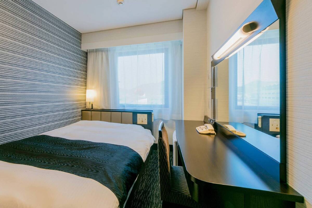 A hotel room with a bed, bedside table, lamp, and a large mirror.