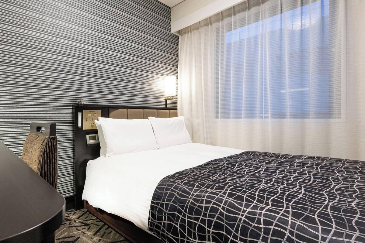 A hotel room with a bed, a bedside table, a lamp, and a window with curtains.