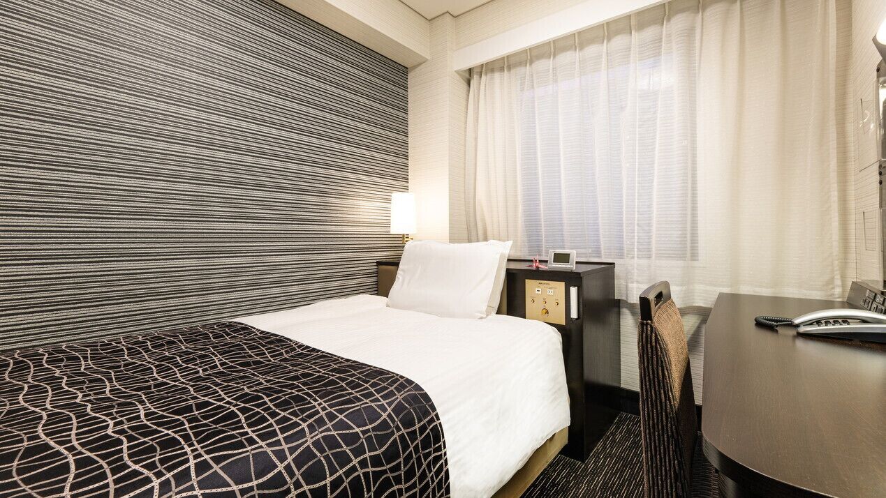 A hotel room with a bed, a desk, a chair, a nightstand, and a window with curtains.