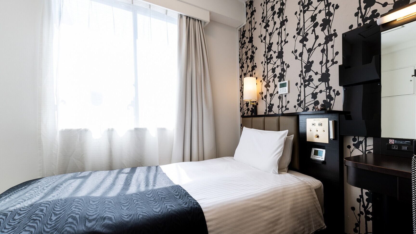 A hotel room with a bed, a bedside table, a mirror, a wall-mounted control panel, and a window with curtains.