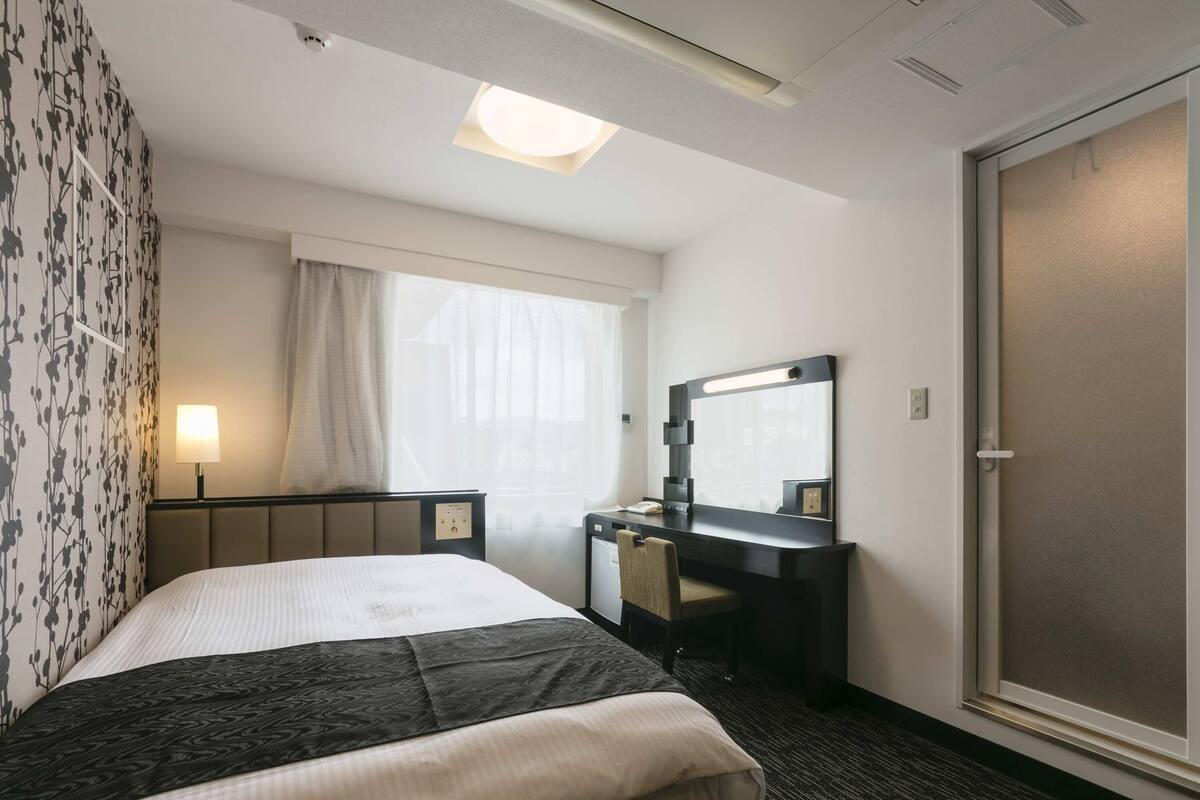 A hotel room with a bed, a desk, a mirror, and a window with curtains.