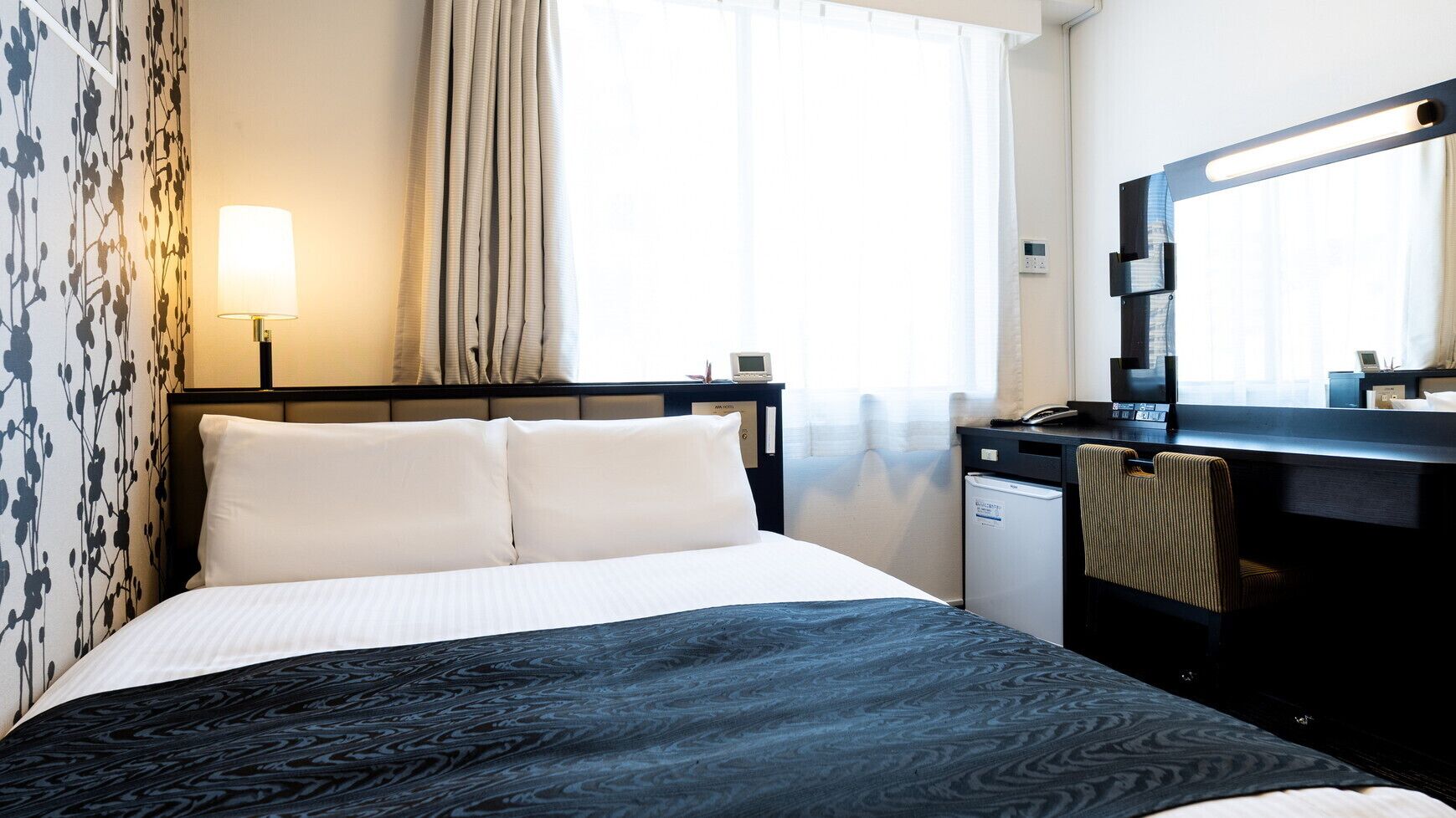 A hotel room with a bed, a desk with a computer, a small refrigerator, and a window with curtains.