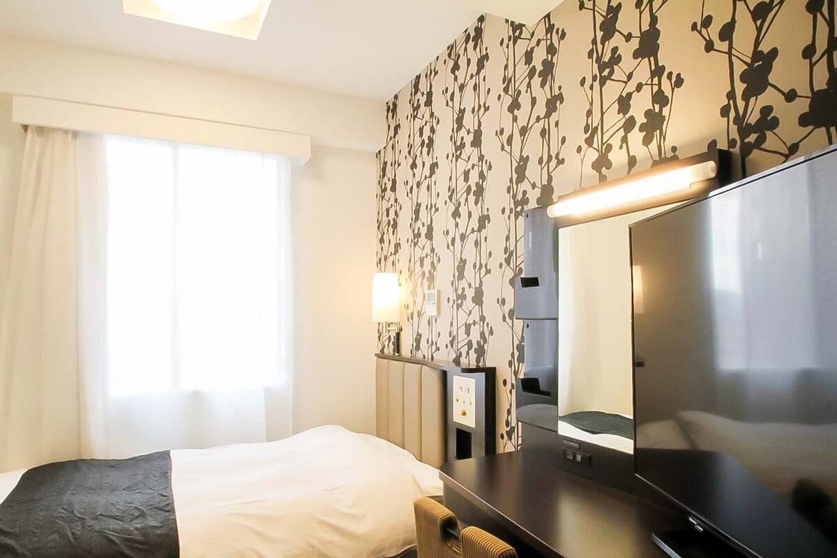 A hotel room with a bed, a desk, a flat-screen TV, a lamp, and a wall with a leaf pattern.