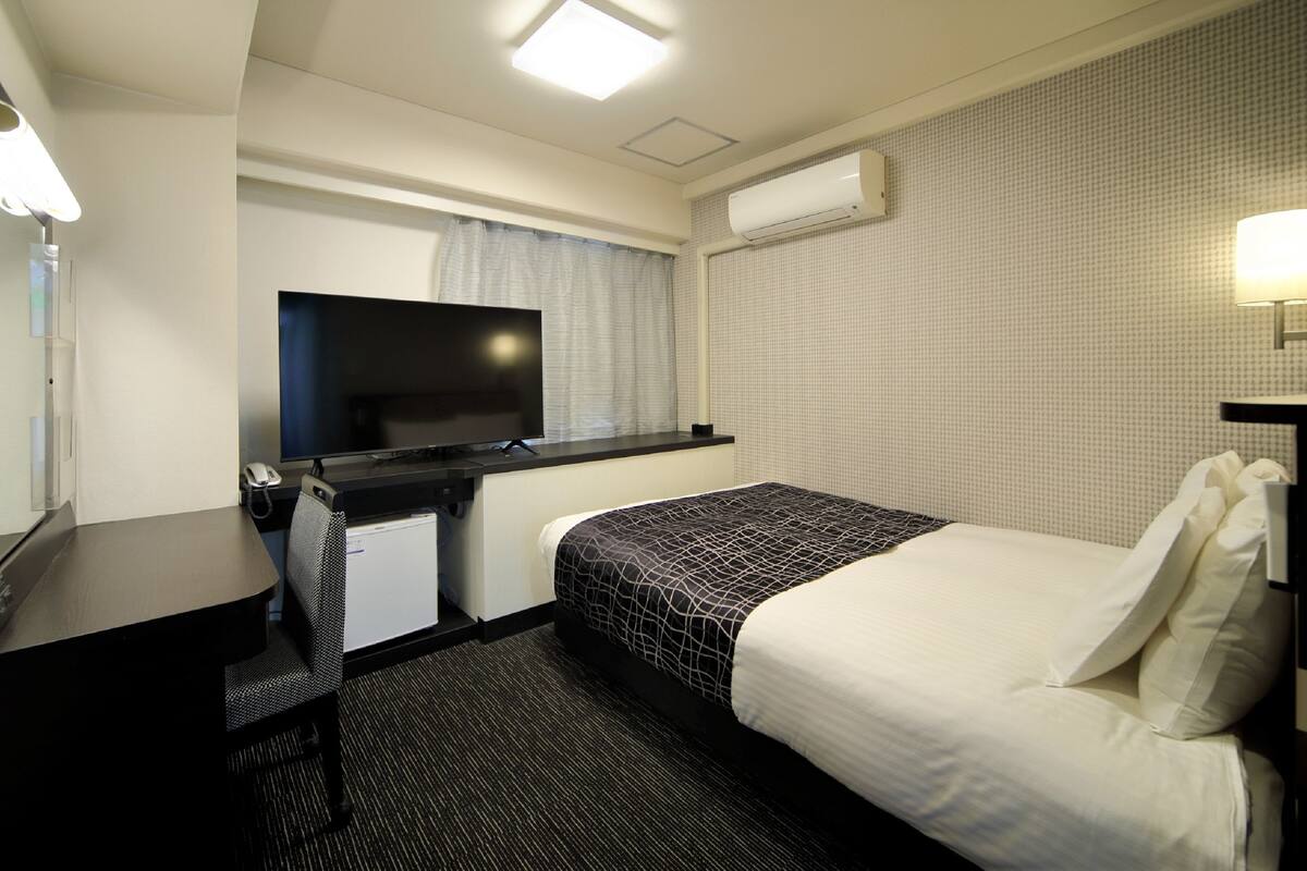 A hotel room with a bed, a desk, a flat-screen TV, an air conditioner, and a window with curtains.