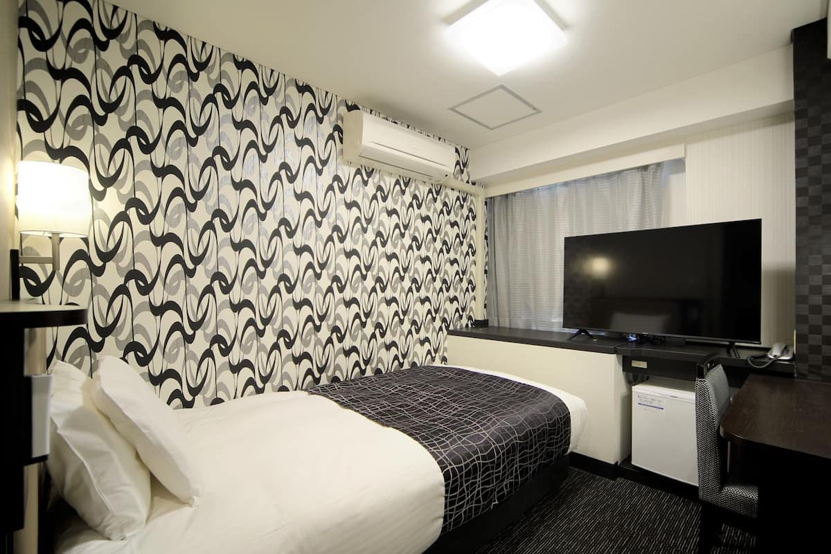 A hotel room with a bed, a desk with a flat-screen TV, a small refrigerator, and a bathroom with a mirror and lighting.