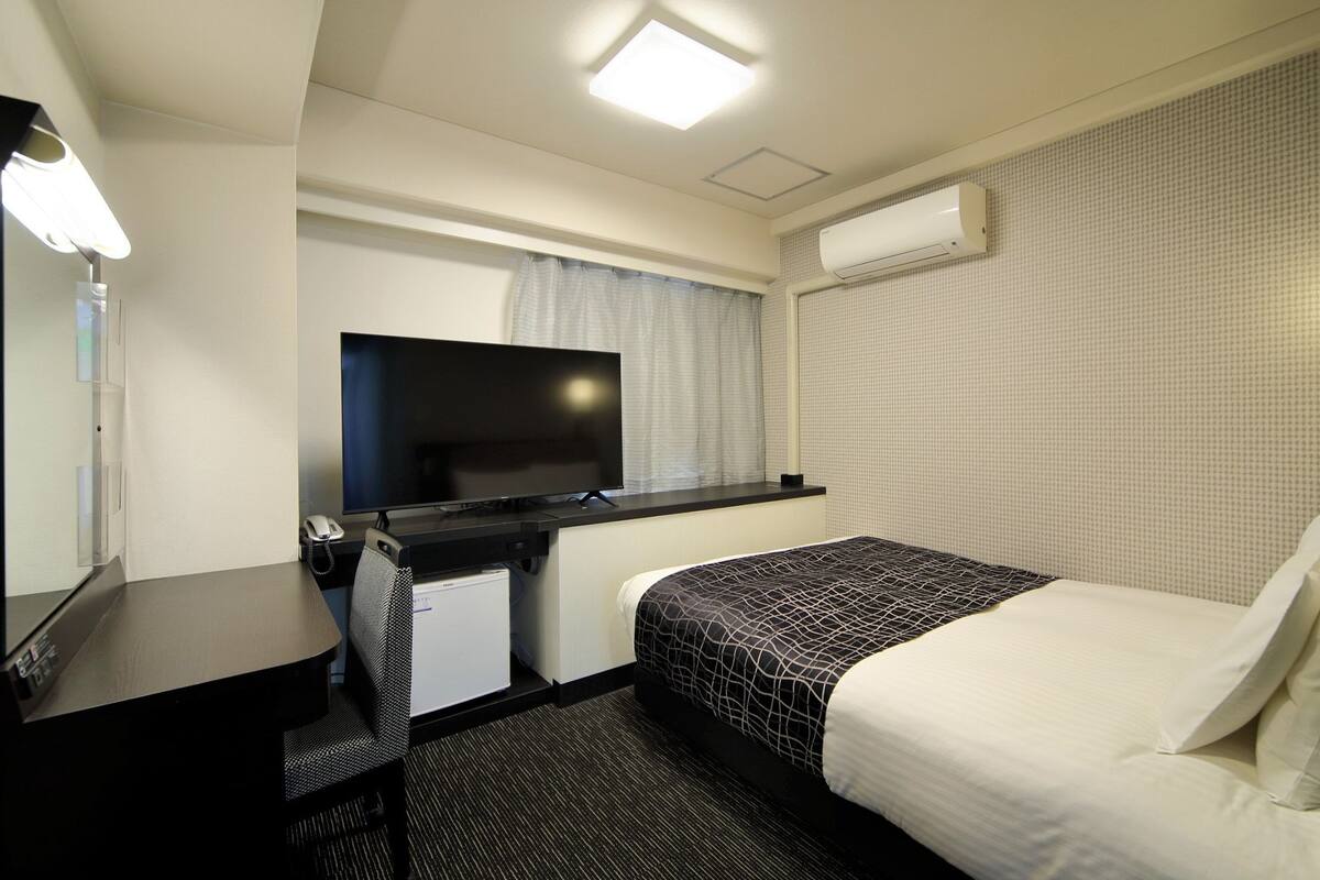 A hotel room with a bed, a desk with a TV, a chair, a desk lamp, and an air conditioning unit.