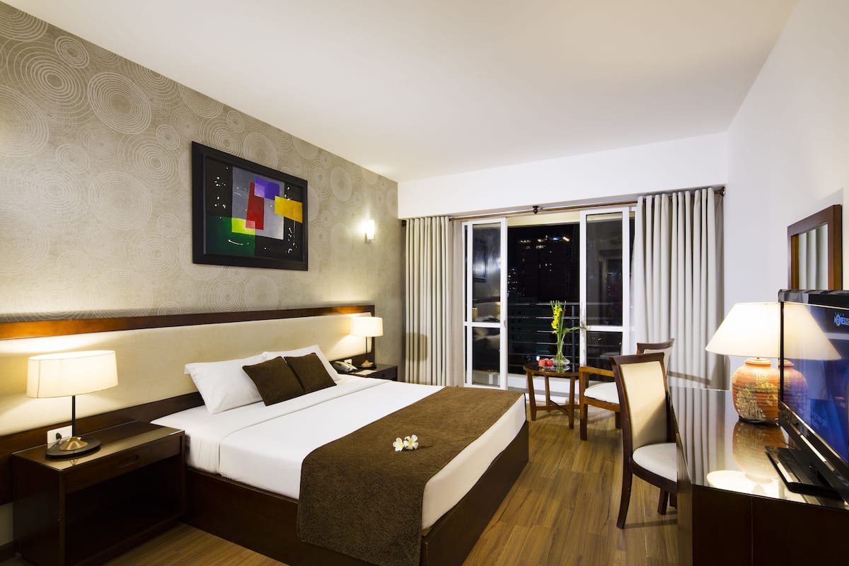 Habitación de hotel con cama, mesitas de noche, escritorio con lámpara, una silla y un gran ventanal con cortinas.