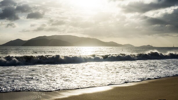 Una playa con olas, una orilla arenosa y montañas a lo lejos.