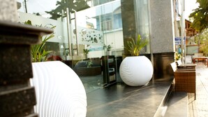 Un hall d’hôtel moderne, agrémenté de grands bacs à plantes blancs, d’un mur de verre qui reflète l’intérieur et d’un espace détente avec des meubles en osier.