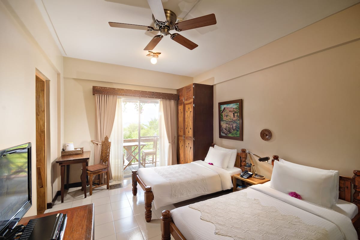 A hotel room with two beds, a desk, a chair, a TV, and a ceiling fan.