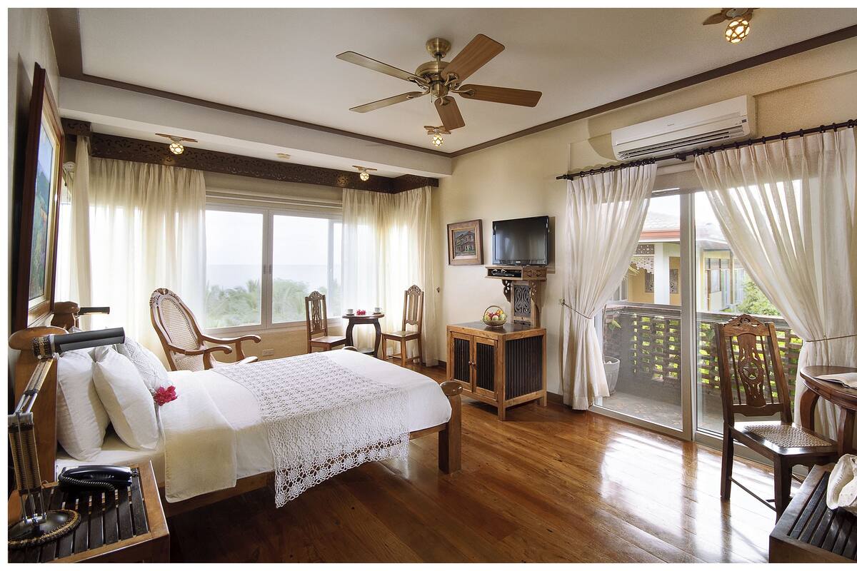 A hotel room with a bed, a television, a desk with a chair, a balcony with a view, and a ceiling fan.