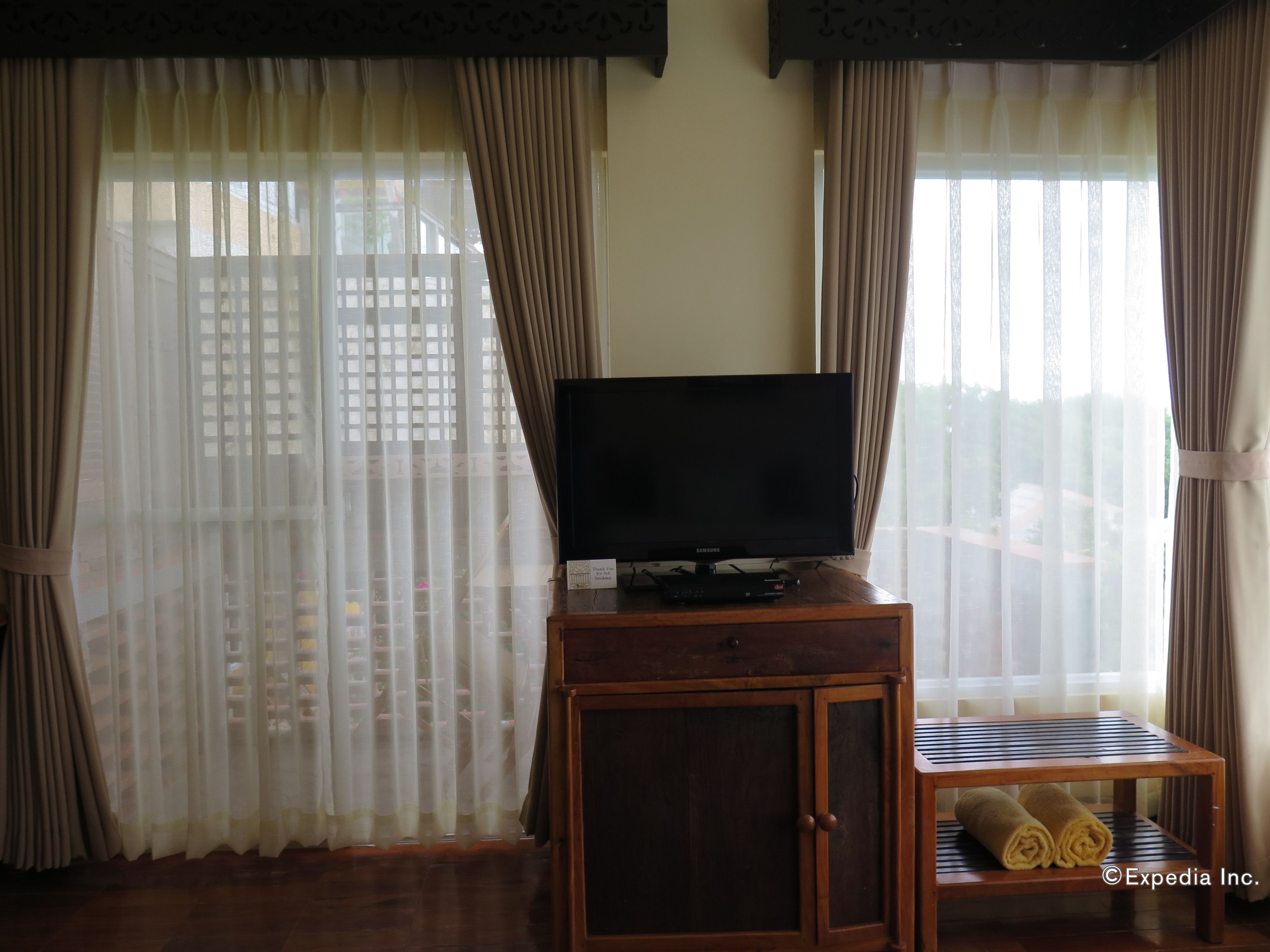 Una habitación con una televisión sobre un mueble de madera, una mesita con una toalla enrollada y cortinas translúcidas que dan a una vista de un edificio.