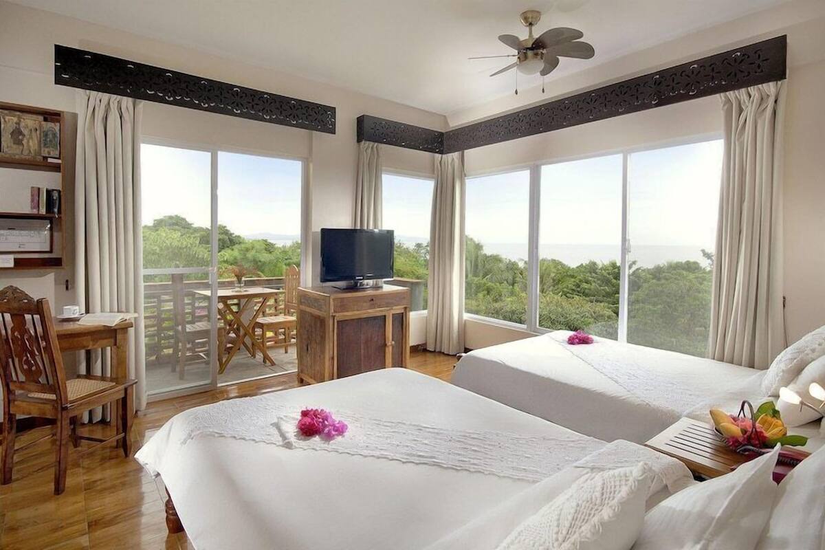 A hotel room with two beds, a desk with a TV, a balcony with a view, and a bookshelf.