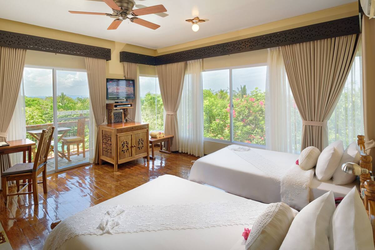 A hotel room with two beds, a TV, a desk, and a view of greenery.