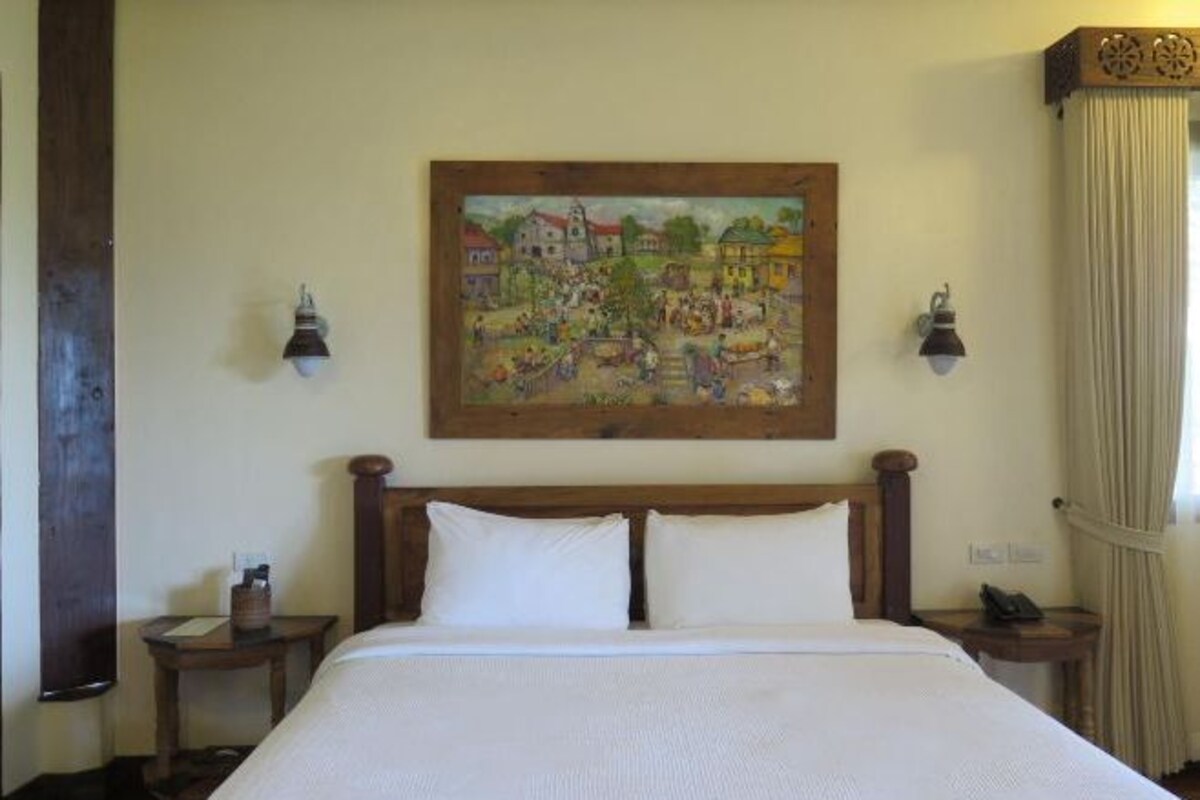Habitación de hotel con una cama grande, cabecera de madera, dos mesitas de noche con lámparas, un cuadro en la pared y una cortina en la ventana.