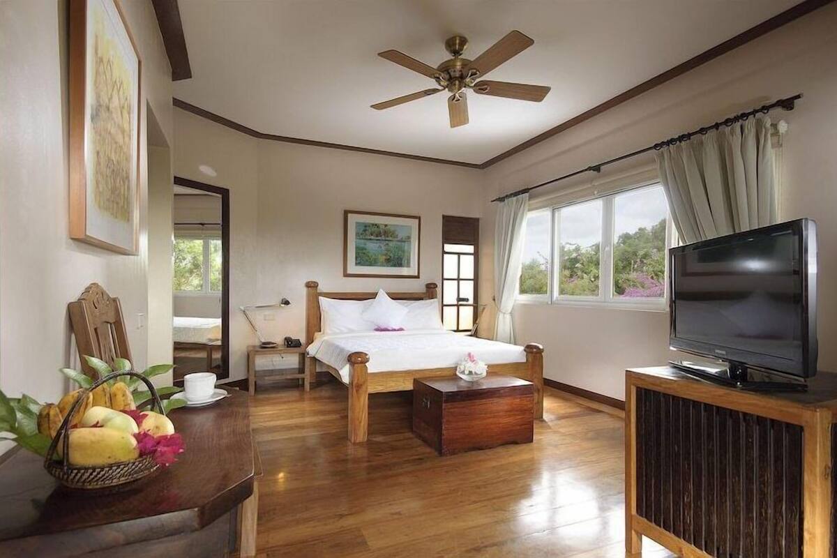 A bedroom with a bed, a television, a ceiling fan, a window with curtains, and a wooden floor.