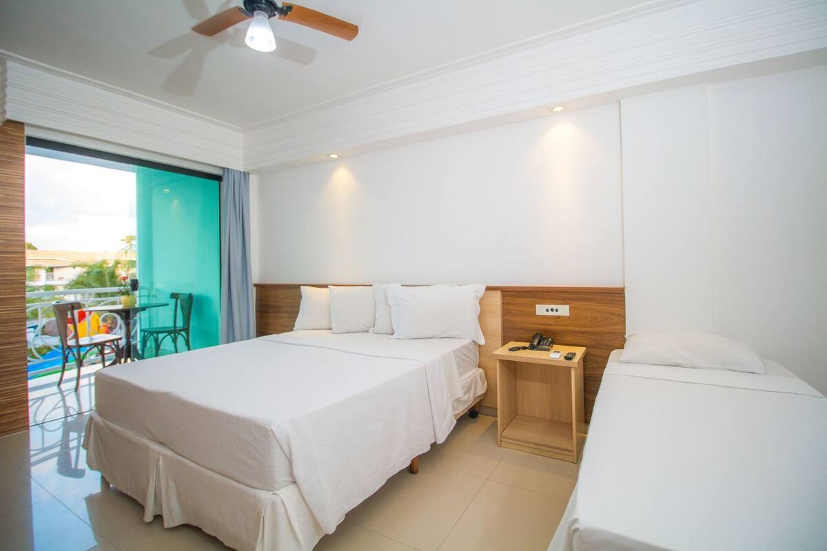 A hotel room with two beds, a wooden nightstand, a ceiling fan, and a view of a balcony with outdoor furniture.