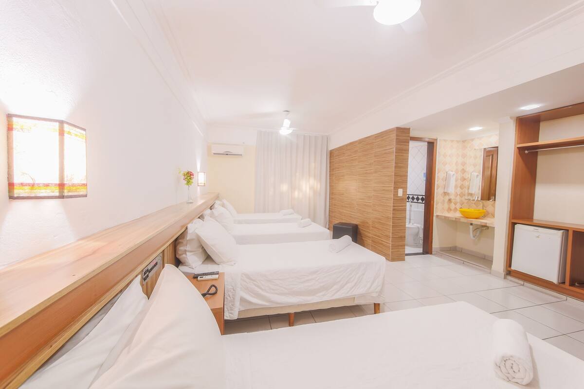 A hotel room with four beds, a wooden headboard, a small table with a remote control, a bathroom with a mirror, and a kitchen area with a sink and a refrigerator.