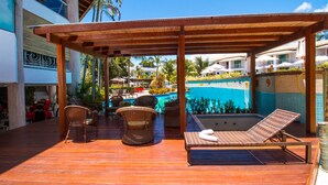A wooden deck with wicker chairs and a lounge chair, overlooking a swimming pool and a building with balconies.