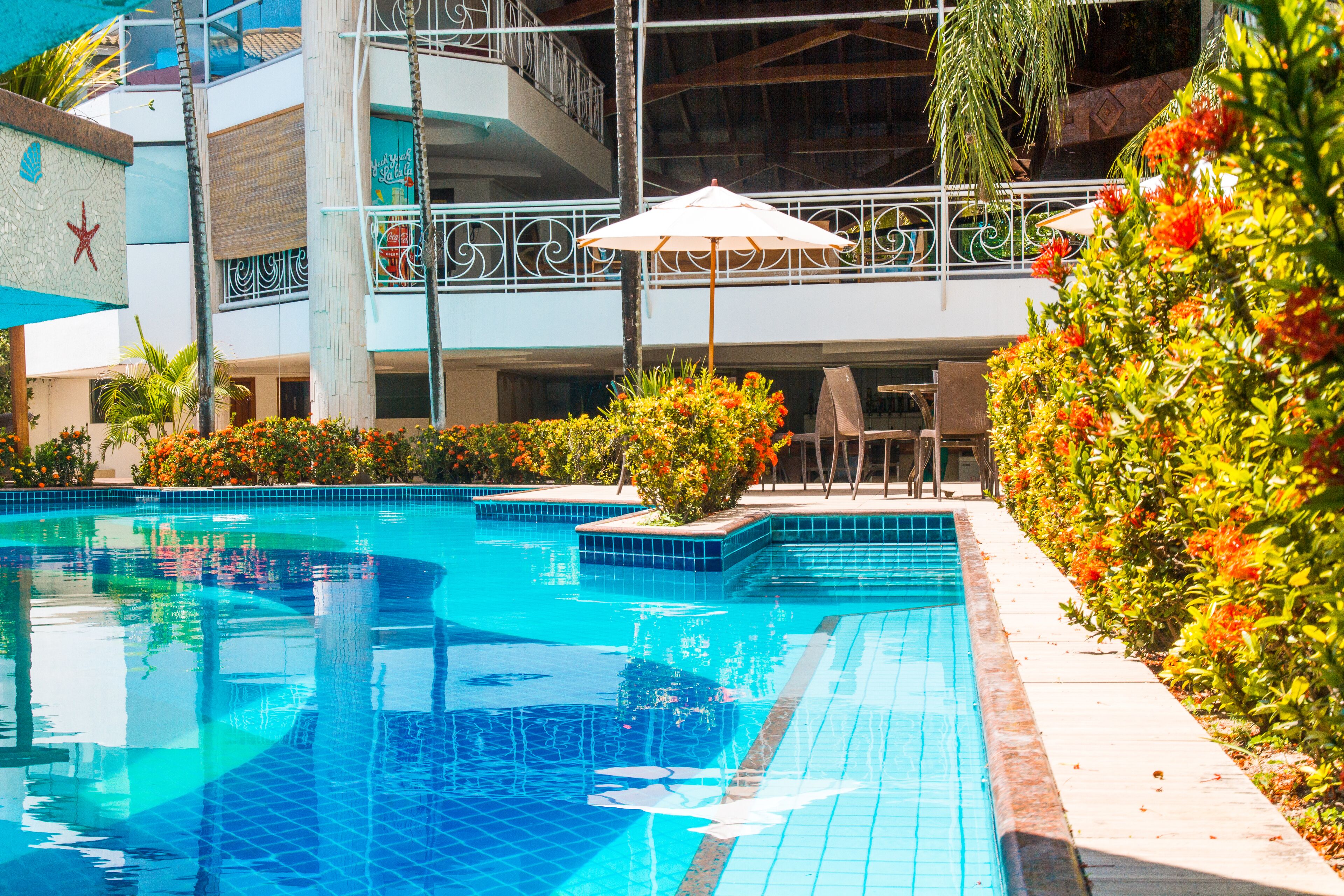 A swimming pool with tiled edges, surrounded by greenery and a building with balconies.