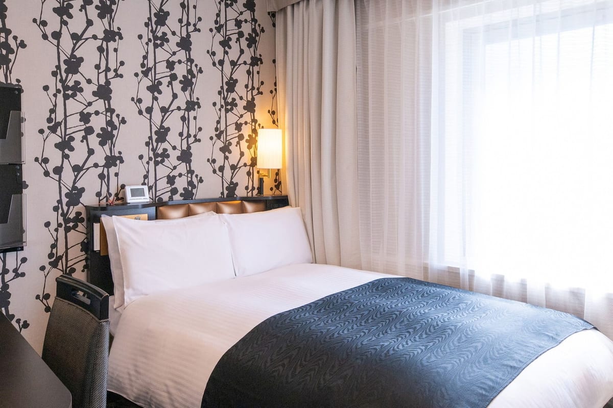 Ein Hotelzimmer mit Bett, Nachttisch, Lampe und einem Fenster mit Vorhängen.