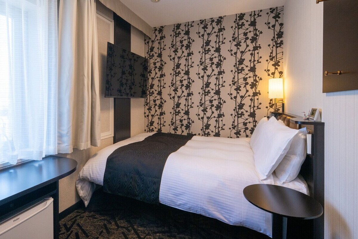 Ein Hotelzimmer mit Bett, Nachttisch, an der Wand montiertem Fernseher und einem Fenster mit Vorhängen.