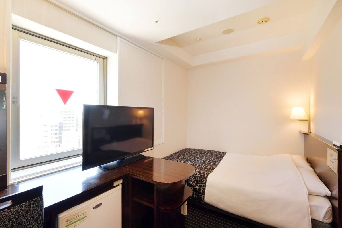 Ein Hotelzimmer mit Bett, Schreibtisch mit Fernseher, kleinem Kühlschrank und Fenster mit Blick auf die Stadt.
