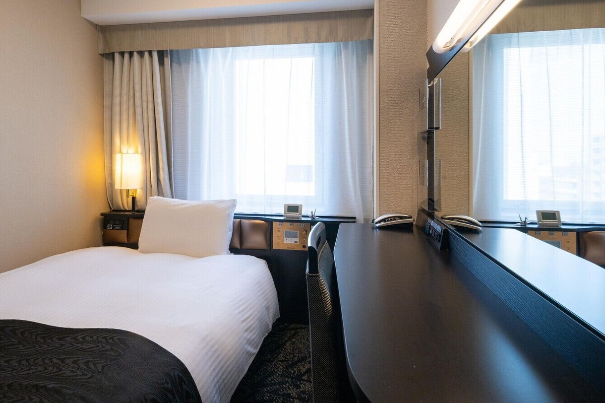 Ein Hotelzimmer mit Bett, Schreibtisch, Stuhl, Lampe und einem großen Fenster mit Vorhängen.