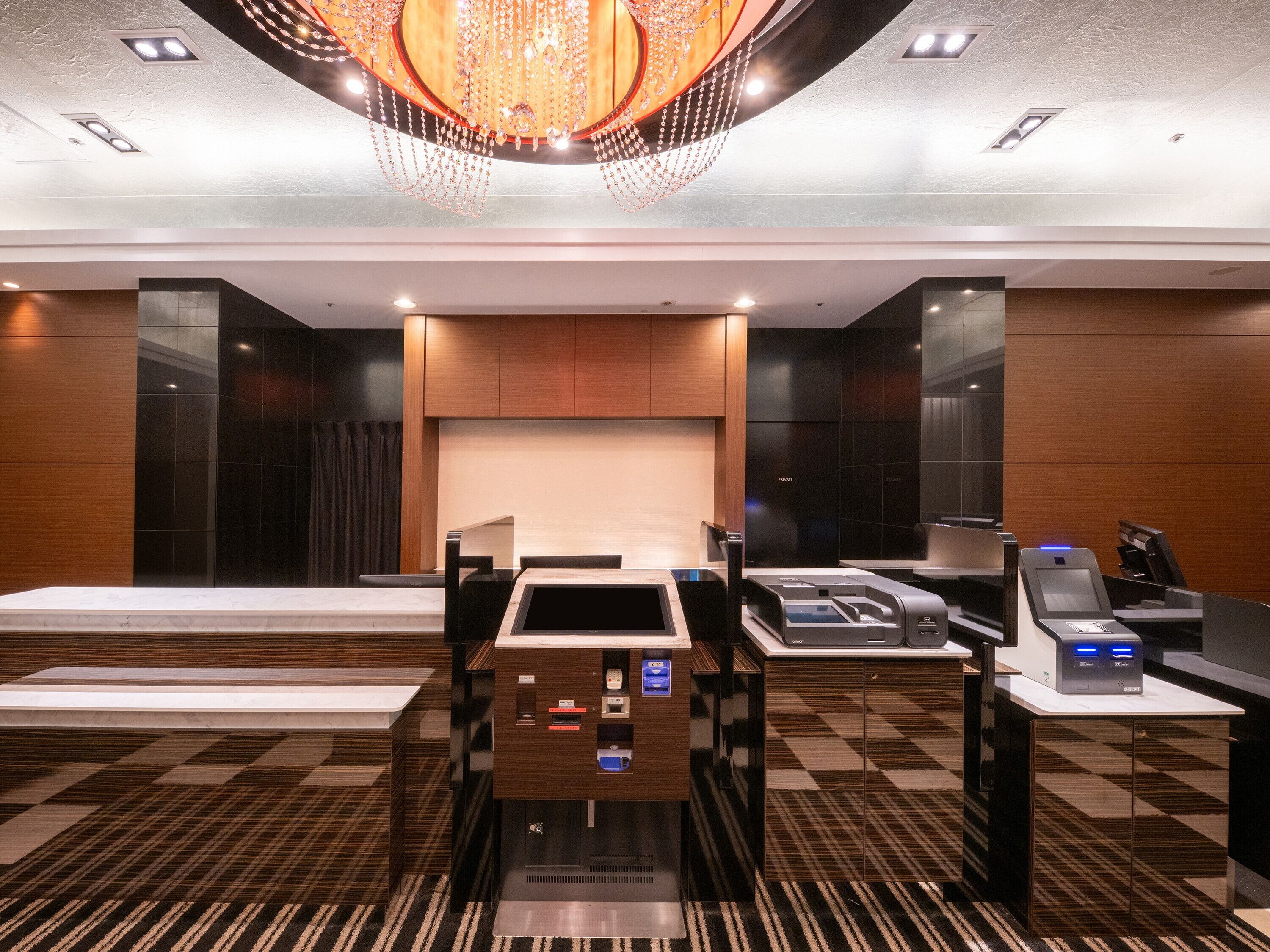 Un vestíbulo de hotel con mostrador de recepción, máquinas de autoservicio, una gran lámpara de araña y un mostrador de check-in.