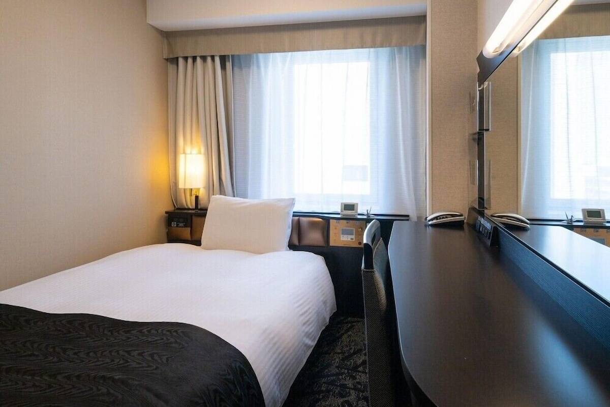 Ein Hotelzimmer mit einem Bett, einem Schreibtisch, einer Lampe, einem Spiegel und einem fenster mit Vorhängen.