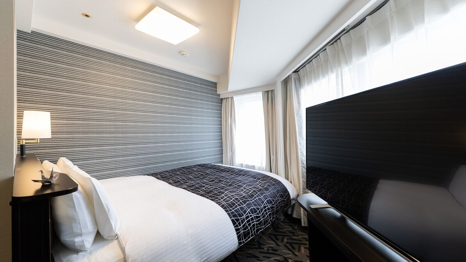 A hotel room with a large bed, a bedside table with a lamp, a flat-screen TV, and a window with curtains.