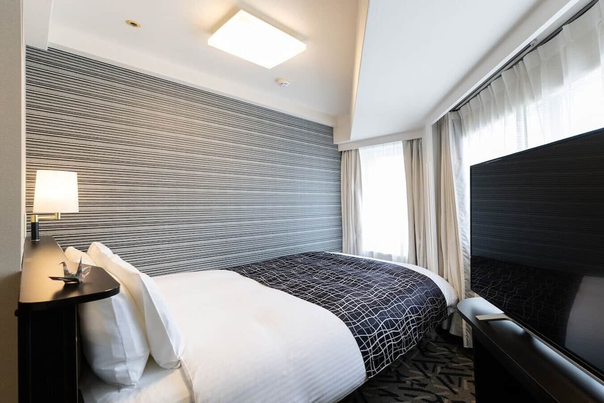 Una habitación de hotel con una cama grande, una mesita de noche con lámpara, un televisor de pantalla plana y una ventana con cortinas.