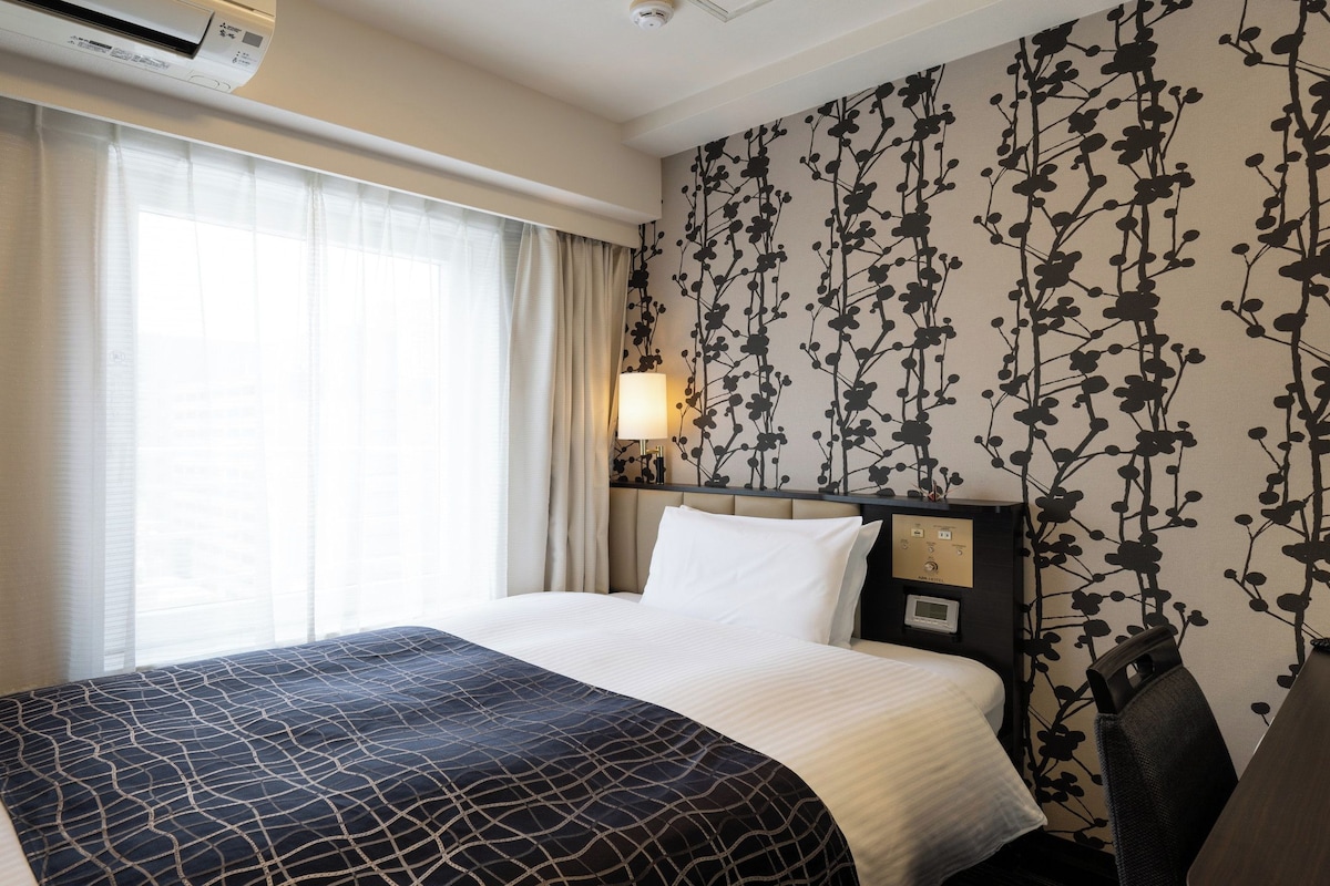 Ein Hotelzimmer mit Bett, Vorhängen, einer Wand-Klimaanlage, einer Nachttischlampe und einem Kopfteil mit Bedienfeld.