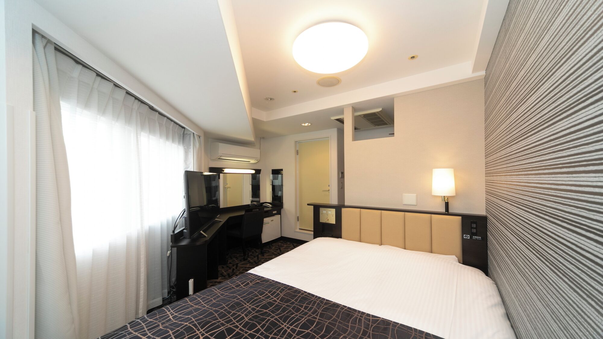 A hotel room with a bed, a desk, a chair, a mirror, a lamp, and a window with curtains.