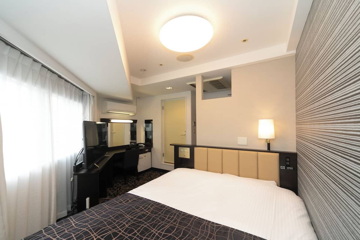 Habitación de hotel con cama, escritorio, silla, espejo, lámpara y ventana con cortinas.
