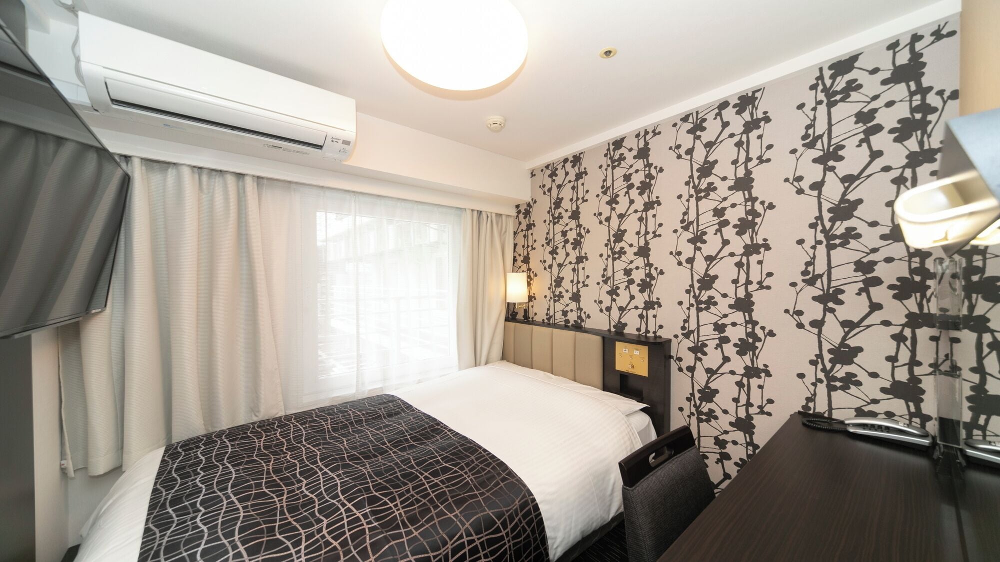 A hotel room with a bed, bedside table, lamp, air conditioning unit, and a window with curtains.