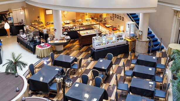 A spacious indoor area with multiple dining tables and chairs, a buffet with various food items, and a seating area with a view of the upper floor.