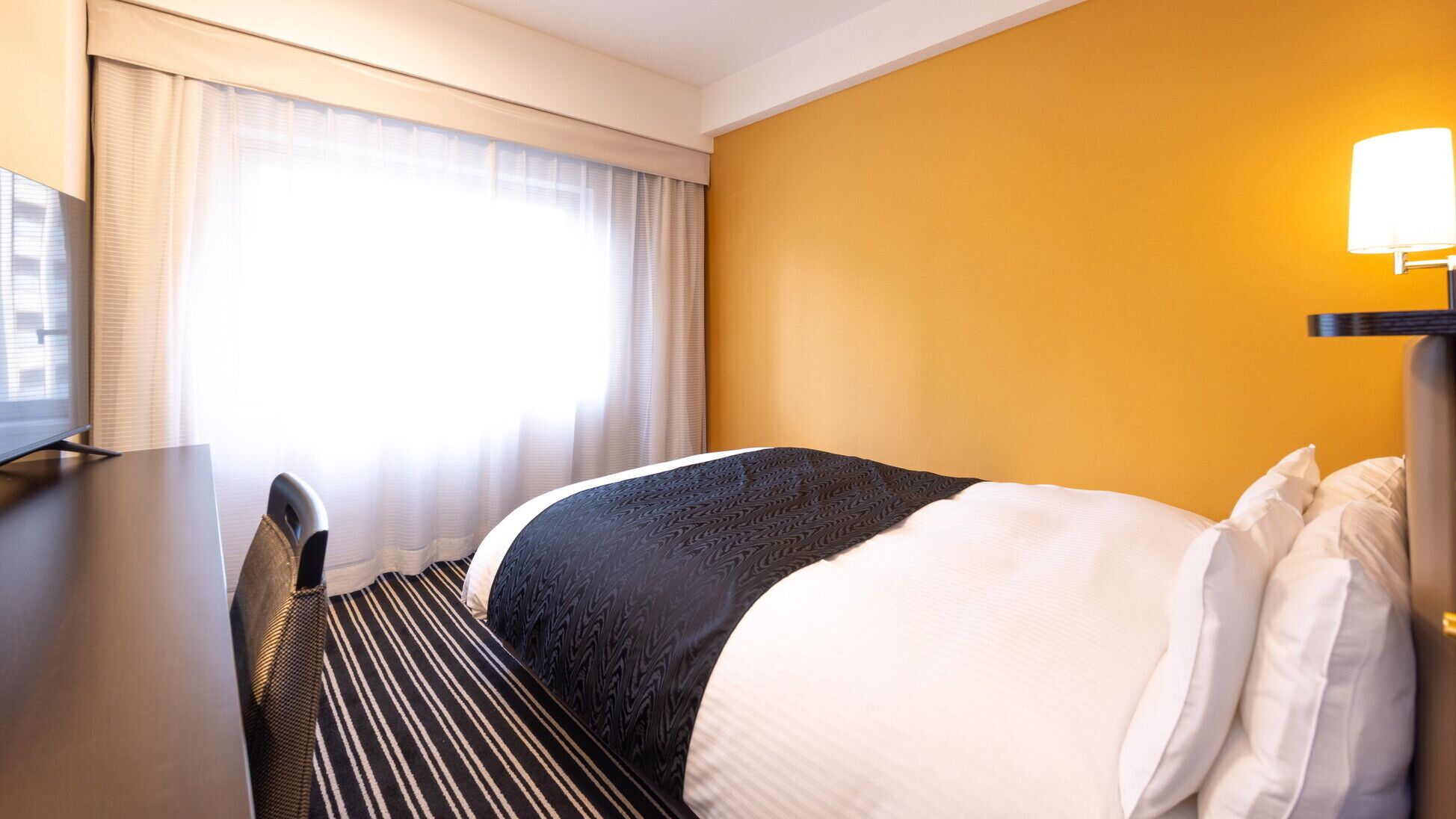 Quarto de hotel com uma cama, uma escrivaninha e uma luminária.