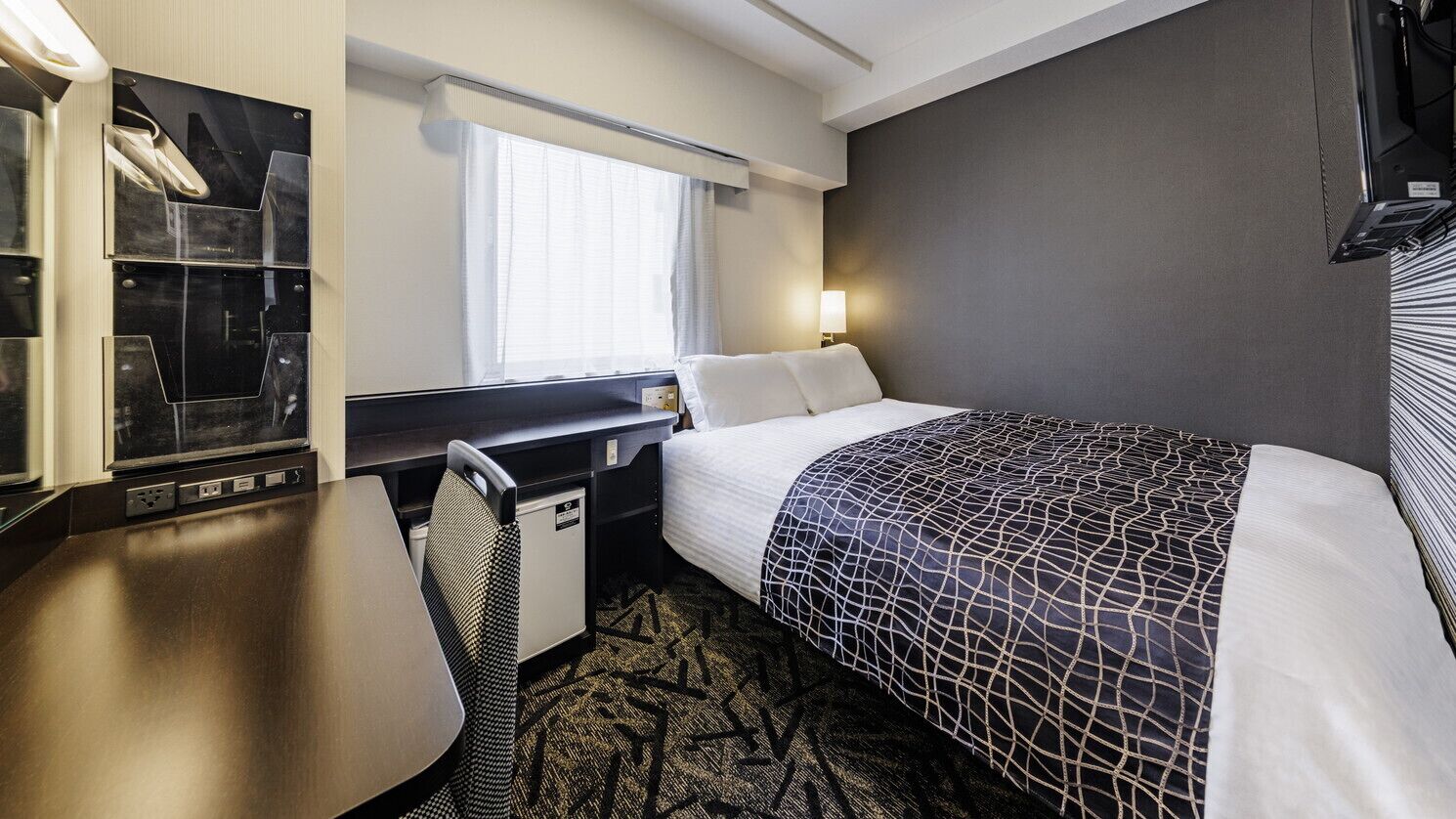 A hotel room with a bed, desk, chair, and a window with curtains.