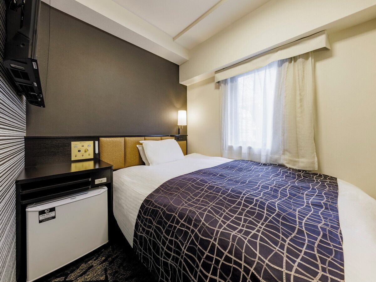 Ein Hotelzimmer mit Bett, Schreibtisch mit Lampe, einem kleinen Kühlschrank und einem Fenster mit Vorhängen.