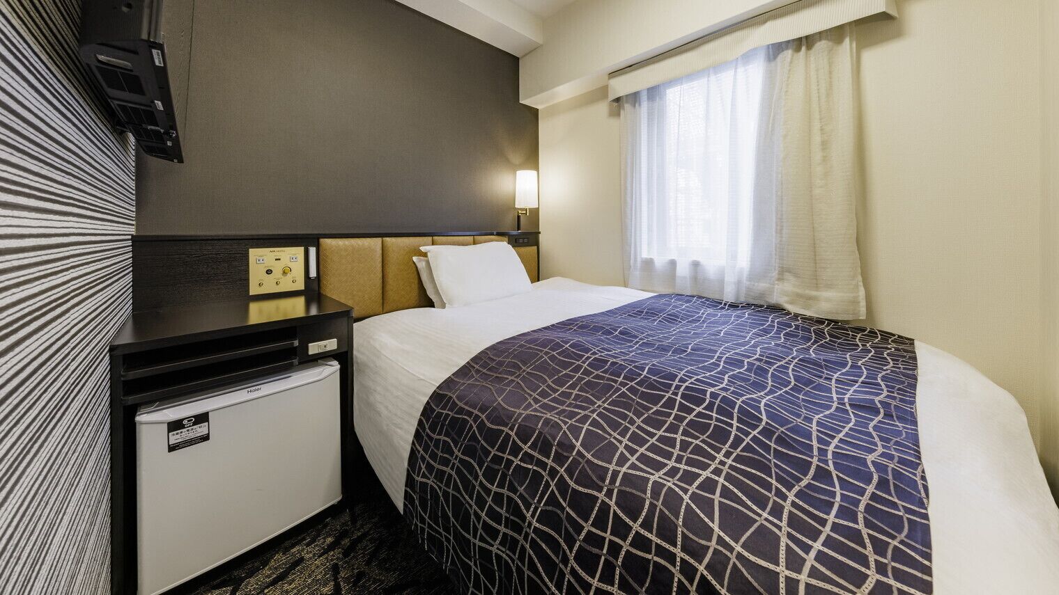 A hotel room with a bed, a nightstand, a small refrigerator, a wall-mounted TV, and a window with curtains.